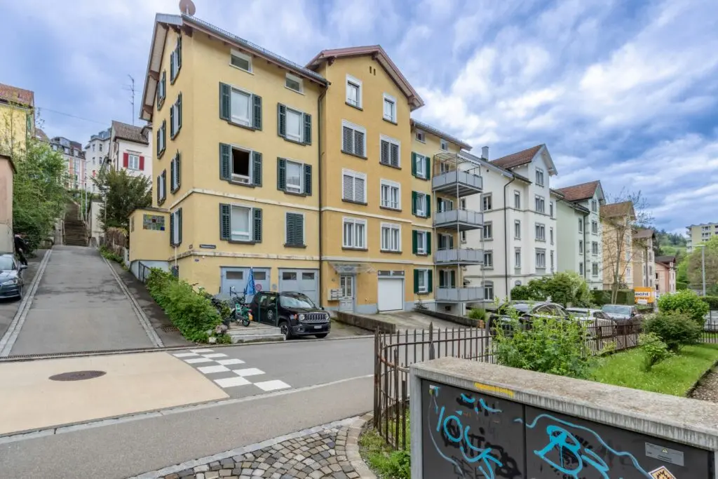 Gelbes Mehrfamilienhaus mit grünen Fensterläden an einer Straßenkreuzung in einer Schweizer Stadt