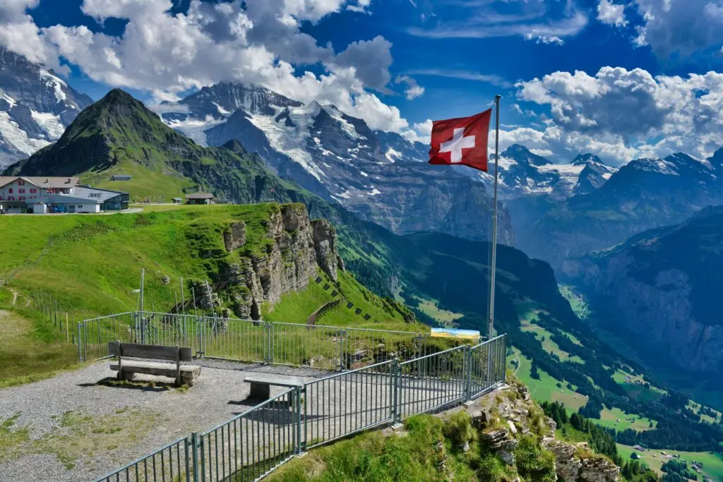 Schweizer Flagge auf einer Aussichtsplattform mit Panoramablick auf schneebedeckte Alpen und grünes Tal