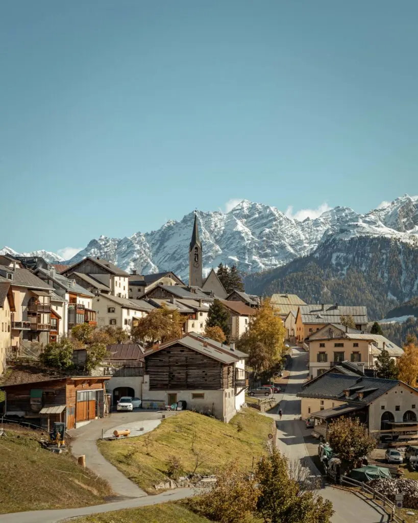 Schweizer Bergdorf mit Kirchturm und schneebedeckten Alpen im Herbst – Graubünden