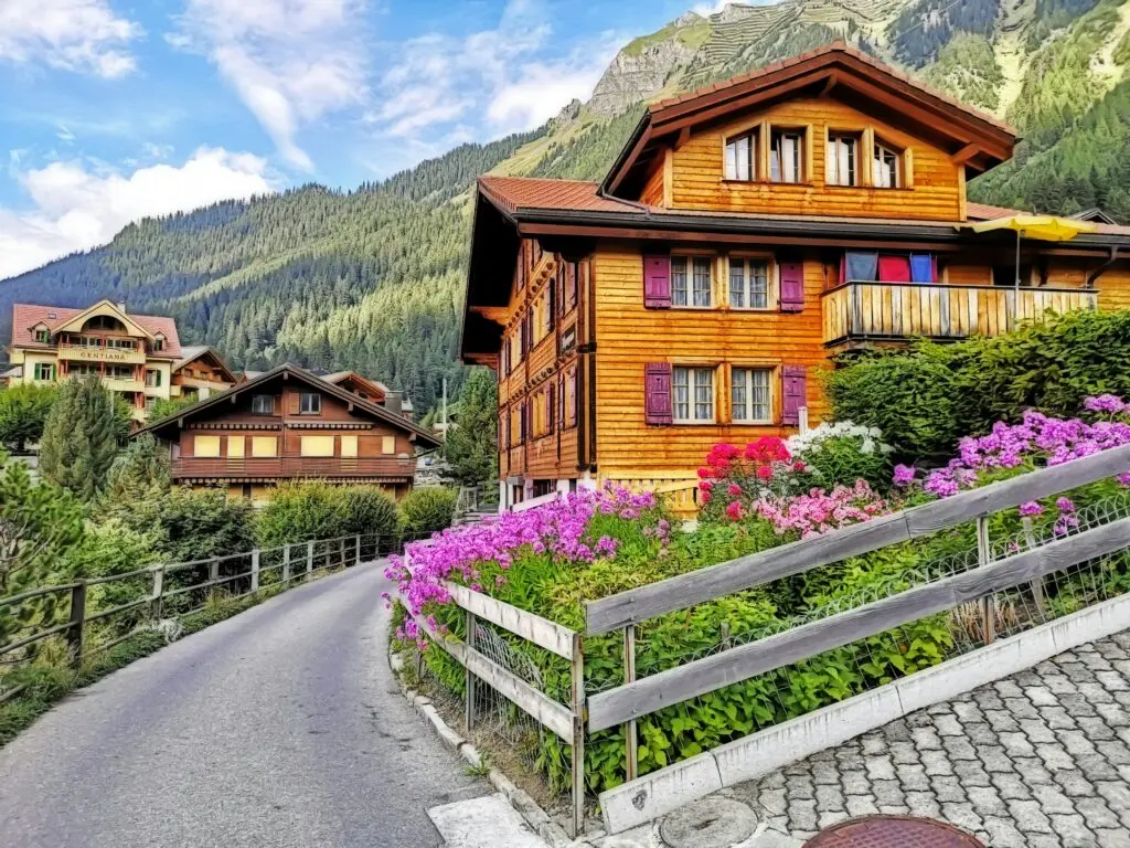 Traditionelles Schweizer Holzchalet mit lila Fensterläden und blühenden Blumen an einer Bergstraße